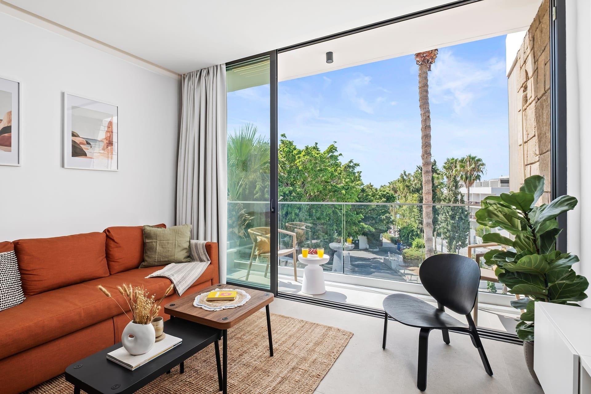 A modern living room with a sofa, coffee table, and chairs, a balcony with a view of greenery and buildings.