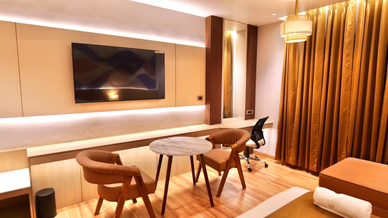 A modern hotel room with a flat-screen TV mounted on the wall, a small round table with two chairs, a sofa, and a large window with curtains.