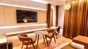 Une chambre d’hôtel moderne dotée d’un téléviseur à écran plat fixé au mur, d’une petite table ronde avec deux chaises, d’un canapé et d’une grande fenêtre avec des rideaux.