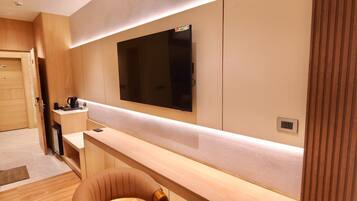 A modern hotel room with a large flat-screen TV mounted on the wall, a wooden desk with a coffee maker, and a comfortable chair.