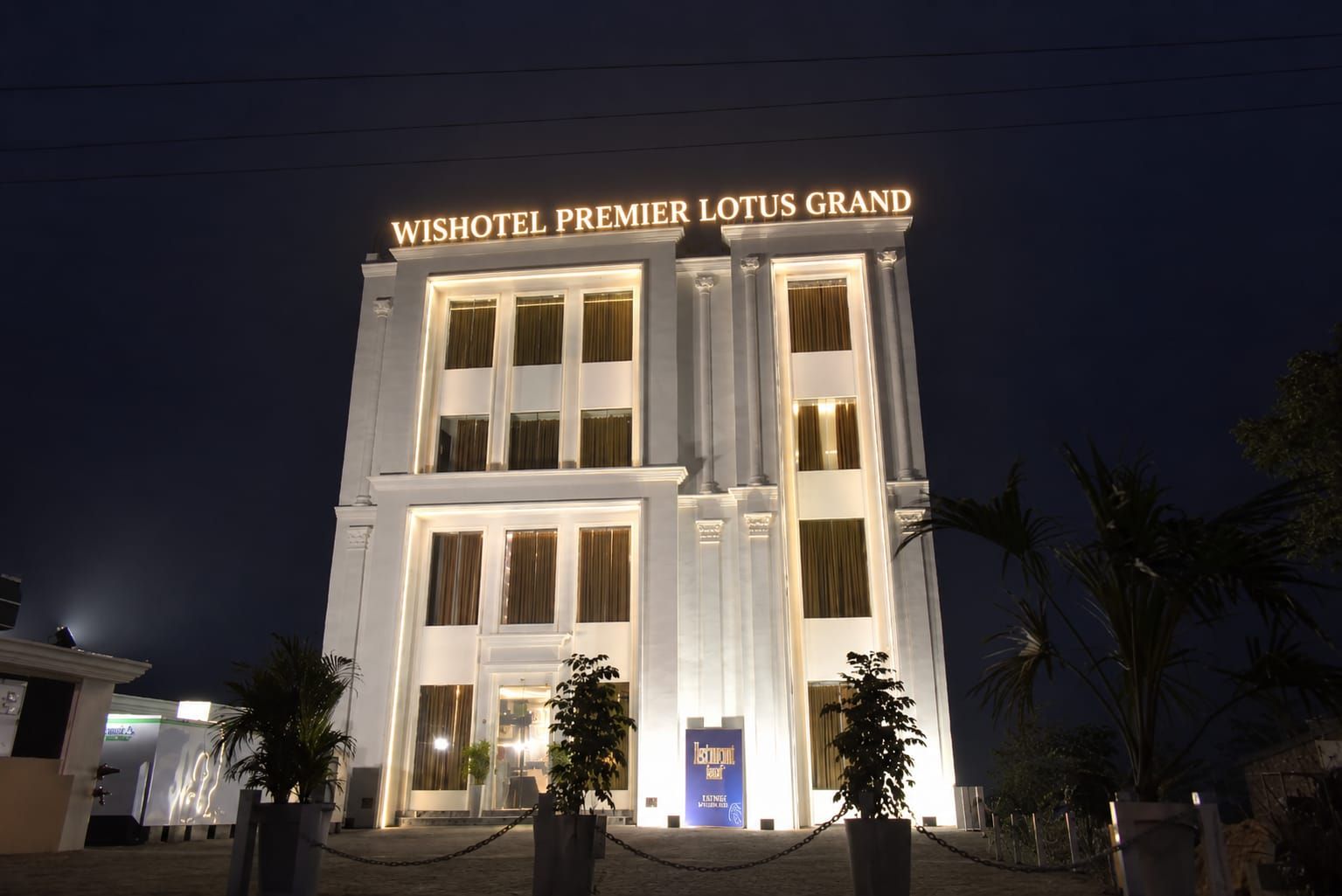 The exterior of the WISHOTEL PREMIER LOTUS GRAND with illuminated signage, large windows, and a symmetrical facade.