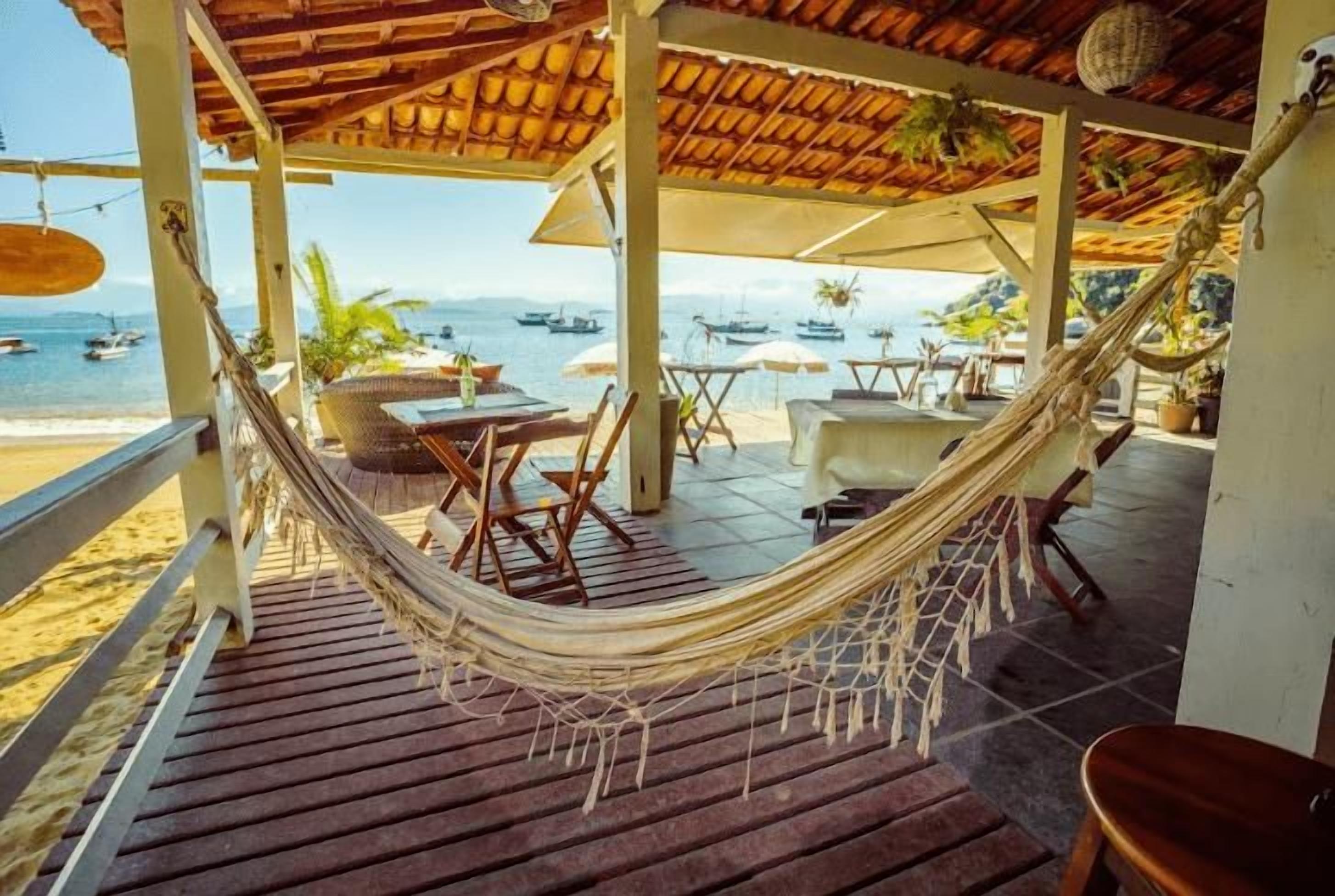 A hammock on a wooden deck overlooking a beach with boats in the distance.