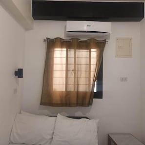 Una habitació amb un llit, una tauleta de nit, un aparell d'aire condicionat i una finestra amb cortina.