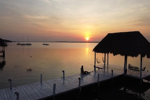 Un atardecer sobre aguas tranquilas, con un muelle y una construcción con techo de paja.