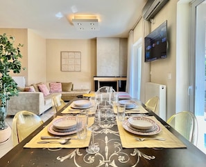 A dining area with a long table set for four, a sofa, a television, and a fireplace. - Hospitality Expert - Pefki Prestige Condo (Pefki)