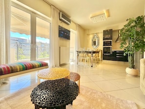 A spacious living area with a large window, a dining table with chairs, and a kitchen area. - Hospitality Expert - Pefki Prestige Condo (Pefki)