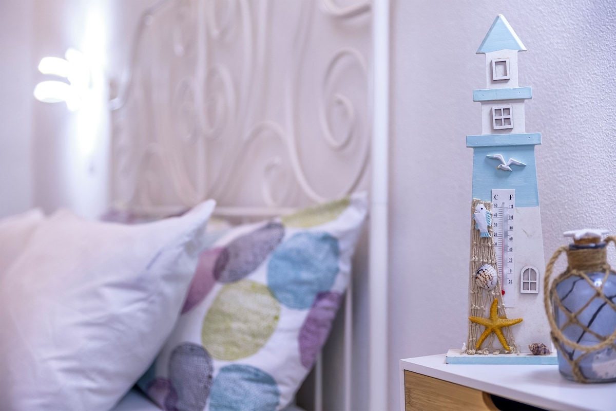 Een bed met een wit hoofdeinde, een decoratief kussen en een klein plankje met een model van een vuurtoren, een zeester en een schelp.