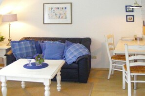 Ein Wohnzimmer mit einem dunkelgrauen Sofa, einem weißen Tisch mit Blumen in einer Vase, einem gerahmten Bild an der Wand und einem Essbereich mit weißen Stühlen und einem Tisch.