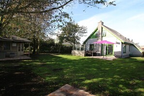 A residential property with a grassy yard, a stone wall, and a house with a green roof and a pink awning. - hollandferien.haus Strandperle 83 (Julianadorp aan zee)