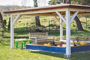Ein überdachter Außenbereich mit Sitzbank und Tisch, umgeben von einem Sandkasten und Spielzeug.