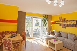 Ein Wohnzimmer mit einer Couch, einem Esstisch und Stühlen. Ein Schiebetürenfenster führt in einen Außenbereich mit Sitzgelegenheiten.