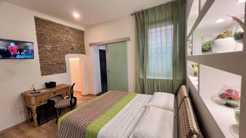 A bedroom with a bed, a desk, a television, a window with curtains, and a wall-mounted shelf with plants.