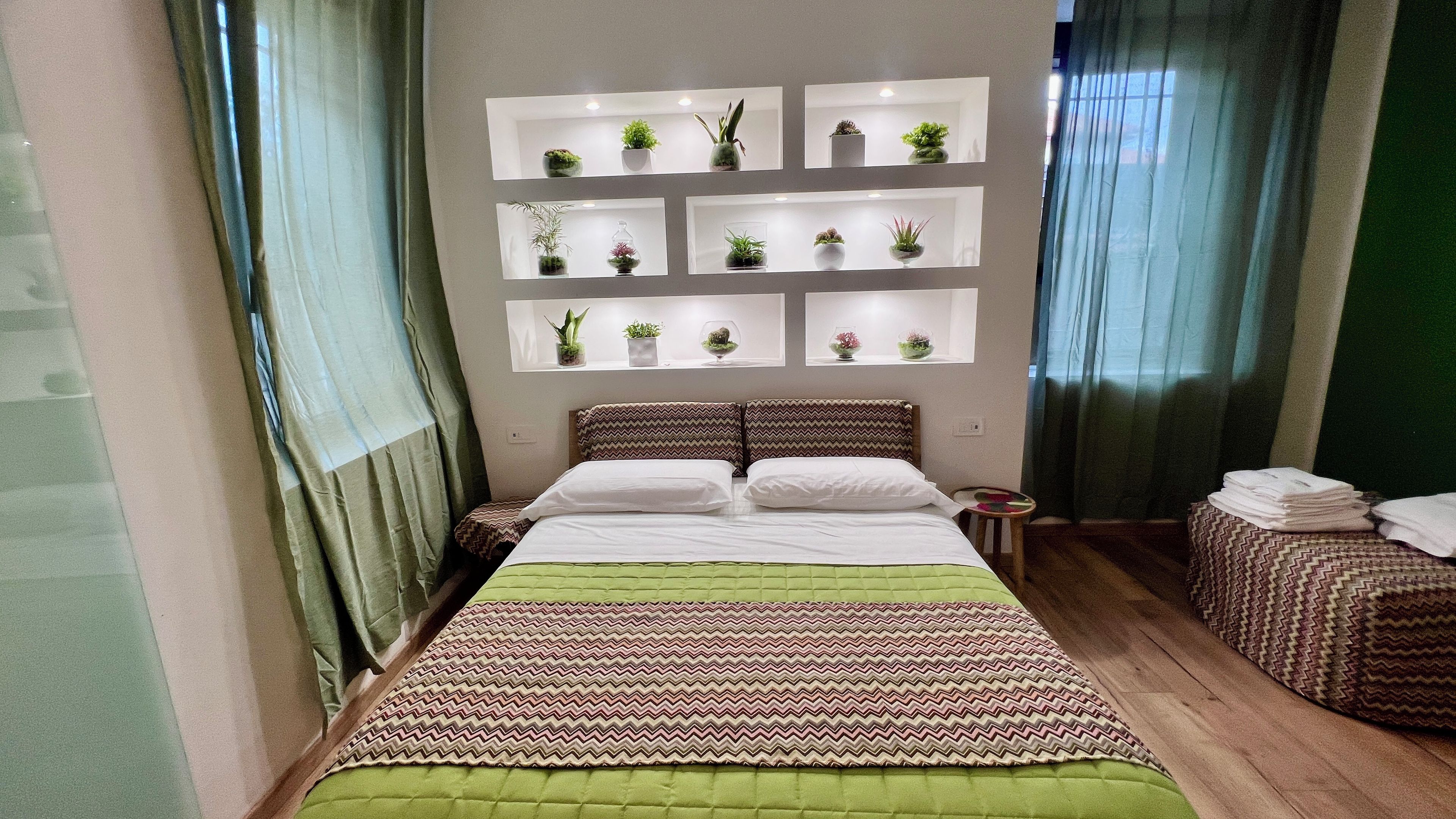 Habitación de hotel con cama, mesita de noche, repisa en la pared con plantas y ventana con cortinas.