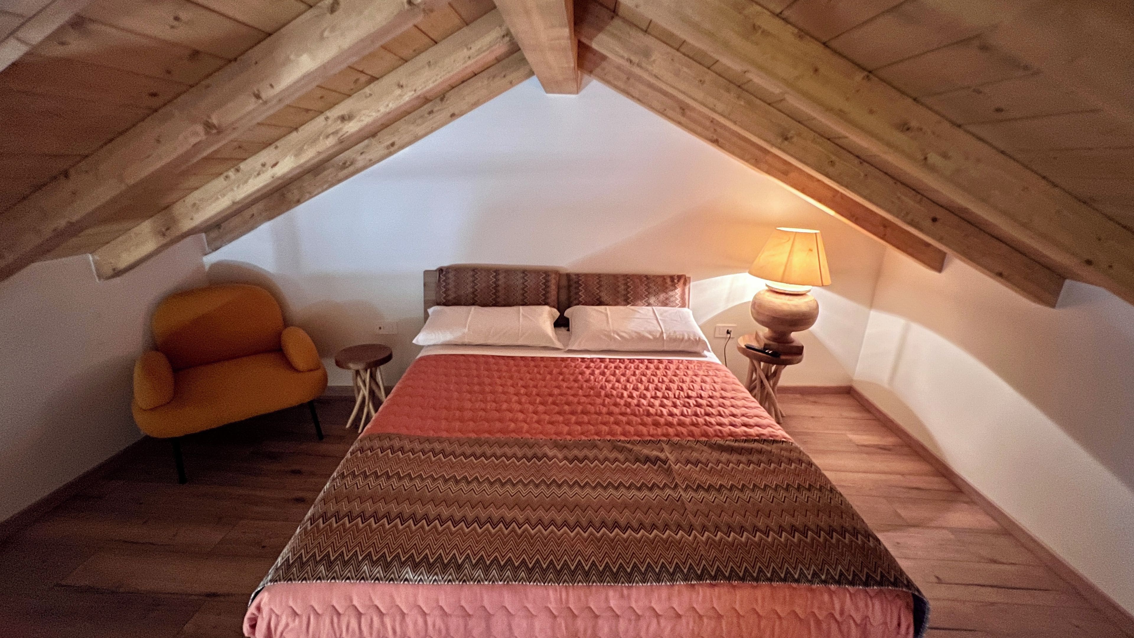 A bedroom with a sloped ceiling, a bed with patterned bedding, a chair, a small table, and a lamp.