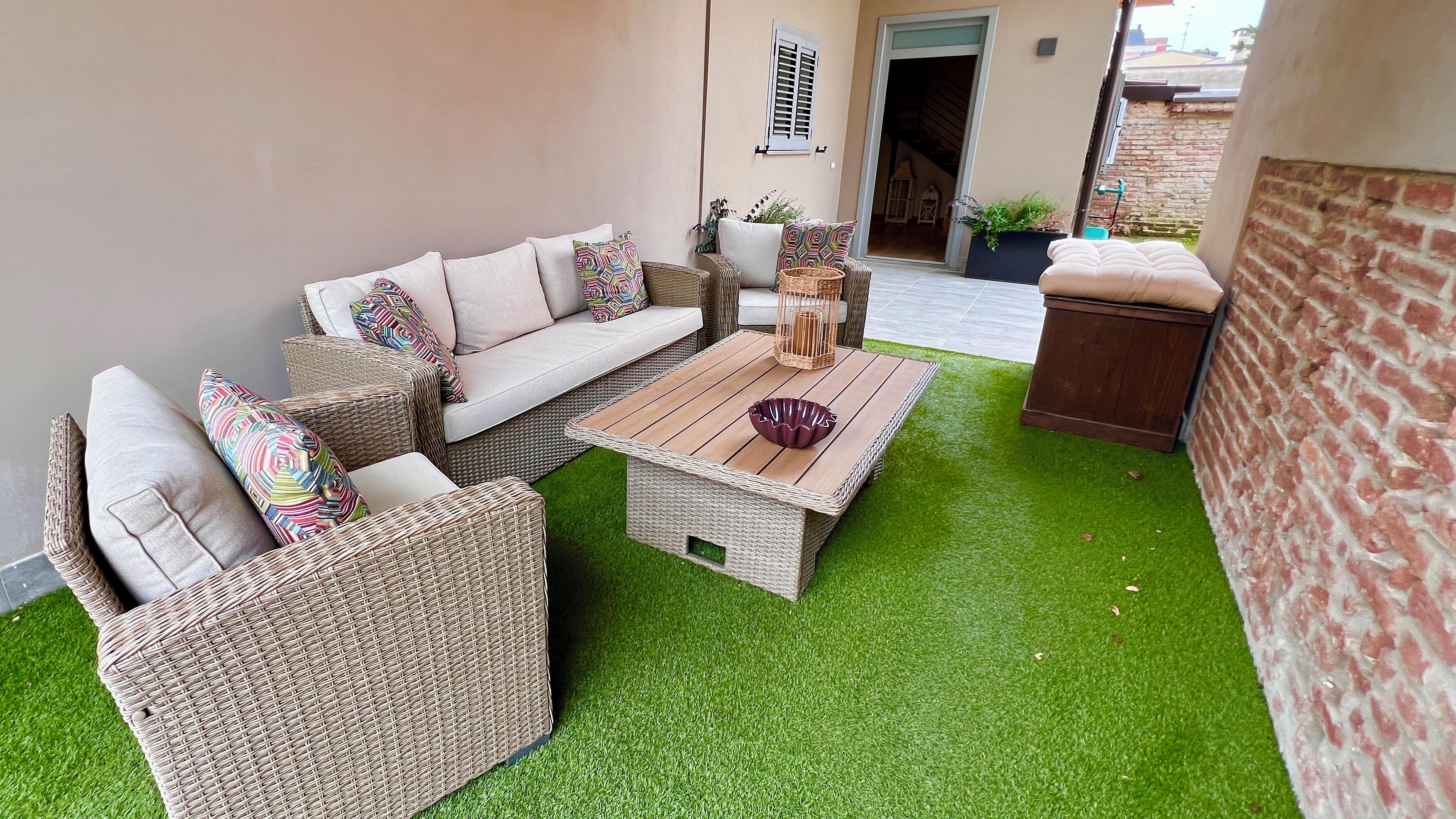Eine Terrassenfläche mit Korbmöbeln, einem Holztisch und Kunstgrase.