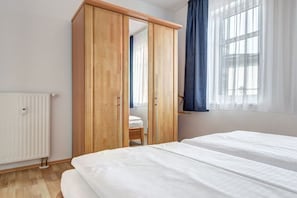 A bedroom with a wooden wardrobe, a white radiator, a window with blue curtains, and two beds. - Villa Sabinium - Apartment 02 (Seebad Heringsdorf)