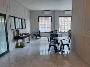 A spacious room with a marble floor, a dining area with tables and chairs, and large windows with blinds. - Ghurfati Hotel Wedana (Jakarta)