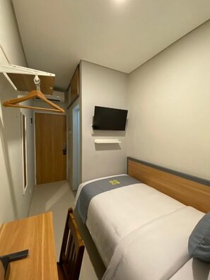 Habitación pequeña con una cama individual, televisión montada en la pared, una silla de madera y una mesita.