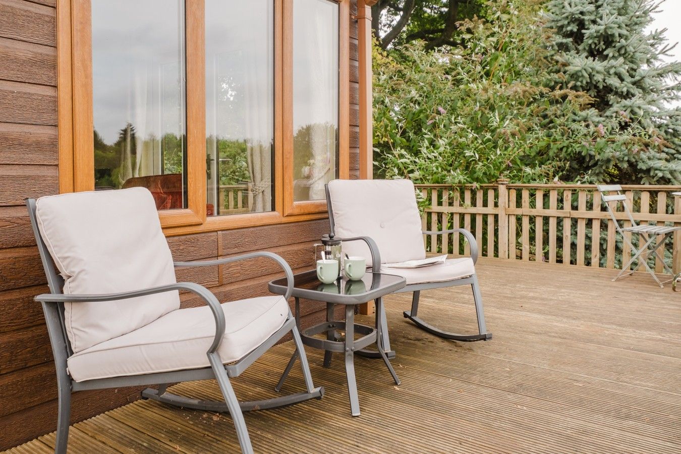 A wooden deck with two rocking chairs and a small table, overlooking a wooden fence and greenery.