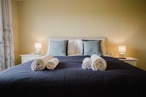 A neatly made bed with rolled towels, a headboard, two bedside tables with lamps, and a floral curtain. - Stunning Lodge For Two in Herefordshire (Holme Lacy)