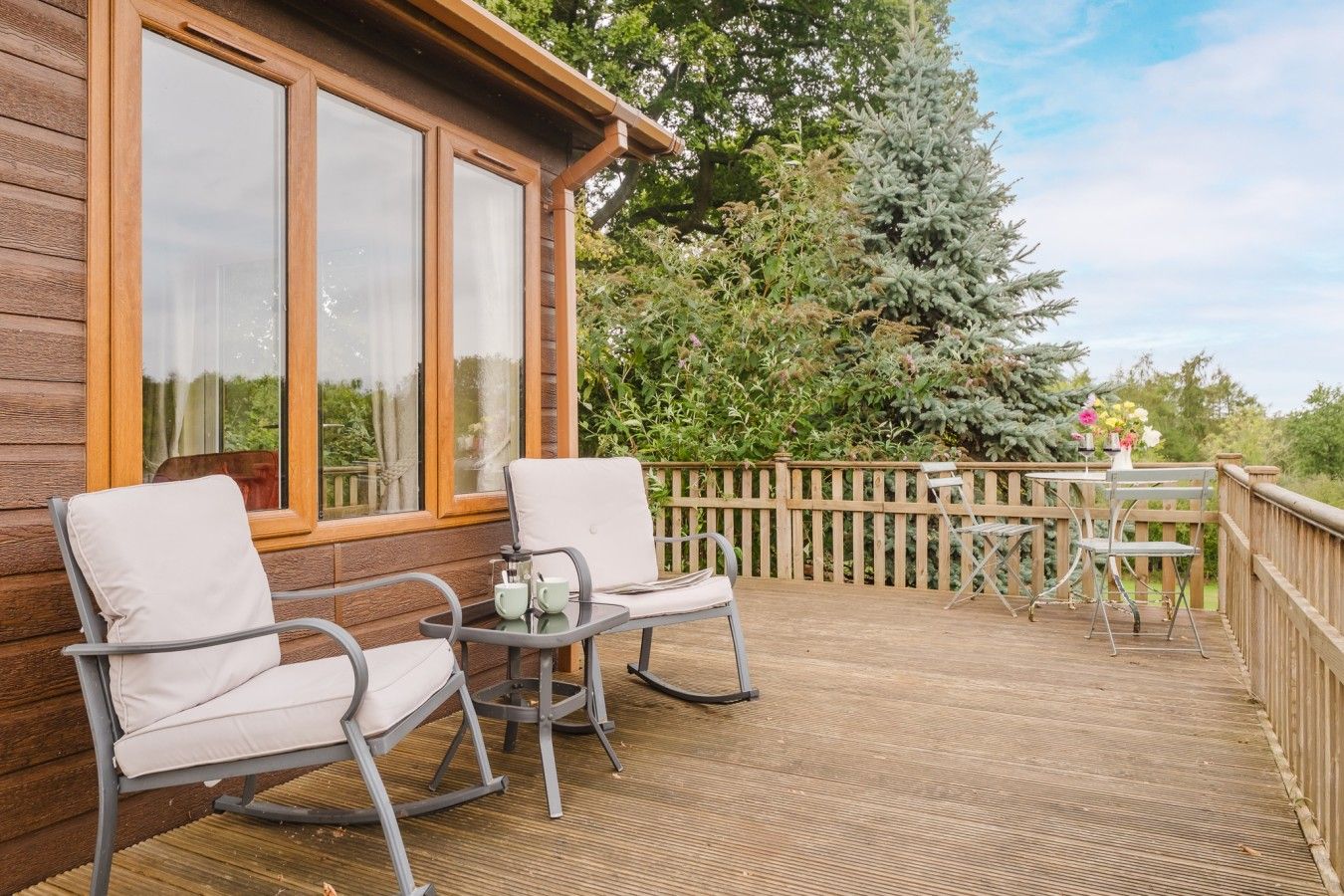 A wooden deck with outdoor seating, including two chairs and a table, and a view of a coniferous tree.