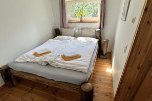 Een enkel bed met houten frame en een grijze matras, een nachtkastje met een lamp en een raam met gordijnen.