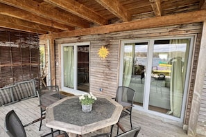 Un patio exterior con terraza de madera, una mesa redonda con sillas y un amplio ventanal corredera.