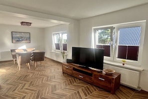 A living room with a herringbone floor, a flat-screen TV on a wooden stand, a dining table with chairs, and large windows with blinds. - Holiday home in Bansin with swimming pool (Bansin)