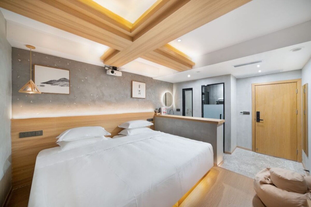 A modern bedroom with a large bed, a wooden ceiling, a bedside table, a mirror, and a door.