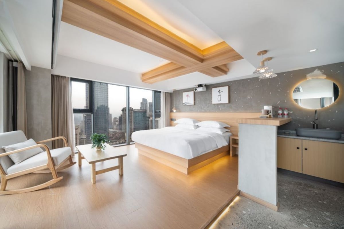 A modern hotel room with a large bed, a desk, a chair, a view of the city, and a small kitchenette.