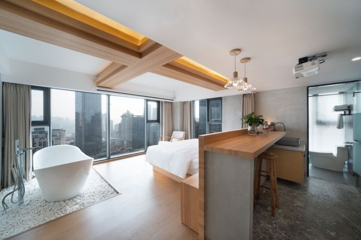 A modern hotel room with a large bed, a bathtub, a kitchen area, and a city view.