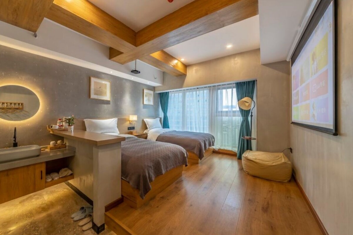 A hotel room with a wooden ceiling, a bed, a bedside table, a mirror, a window with curtains, and a projector screen on the wall.