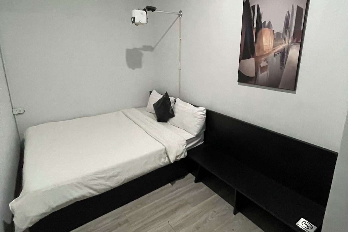 A small, modern bedroom with a single bed, a bedside table, a wall-mounted lamp, and a framed picture on the wall.