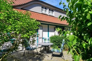 A patio with a round table and two chairs, a building with a red roof, and green foliage in the foreground. - Holiday apartment with 1 bedroom (Dranske)