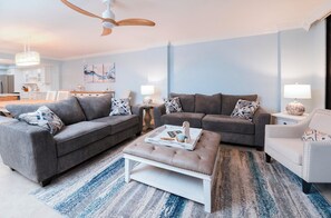 A modern living room with two grey sofas, a white ottoman, and a white armchair. There is a ceiling fan, a chandelier, and a dining area in the background. - #102 Seychelles 2/2 Oceanview Traffic Free Beach (Daytona Beach Shores)