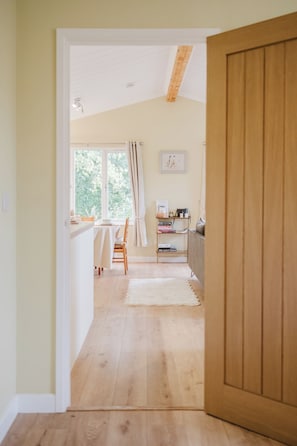 Un pasillo con piso de madera que conduce a un comedor con mesa y sillas, una estantería y una ventana con cortinas.