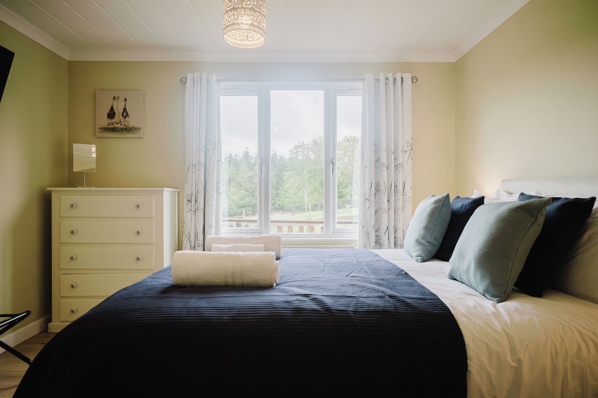 Ein Schlafzimmer mit einem großen Bett, einem Schminktisch, einem Fenster mit Vorhängen und einer Pendelleuchte.