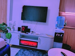 A room with a flat-screen TV mounted on the wall, a water dispenser, a small cabinet, and a plant. - Cozy Studio with Gym & Lift Prime location near TRM Mall (Nairobi)