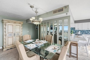 A dining room with a glass table, beige chairs, a chandelier, and a view of the kitchen through a mirrored wall. - 11D Royal Atlantic 2/2 ocean view unit (Daytona Beach Shores)