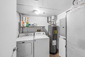 Una sala de bugaderia amb rentadora, assecadora i productes de neteja apilats.