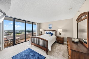 A bedroom with a large bed, a dresser, a nightstand, and a view of the city through floor-to-ceiling windows. - 11D Royal Atlantic 2/2 ocean view unit (Daytona Beach Shores)