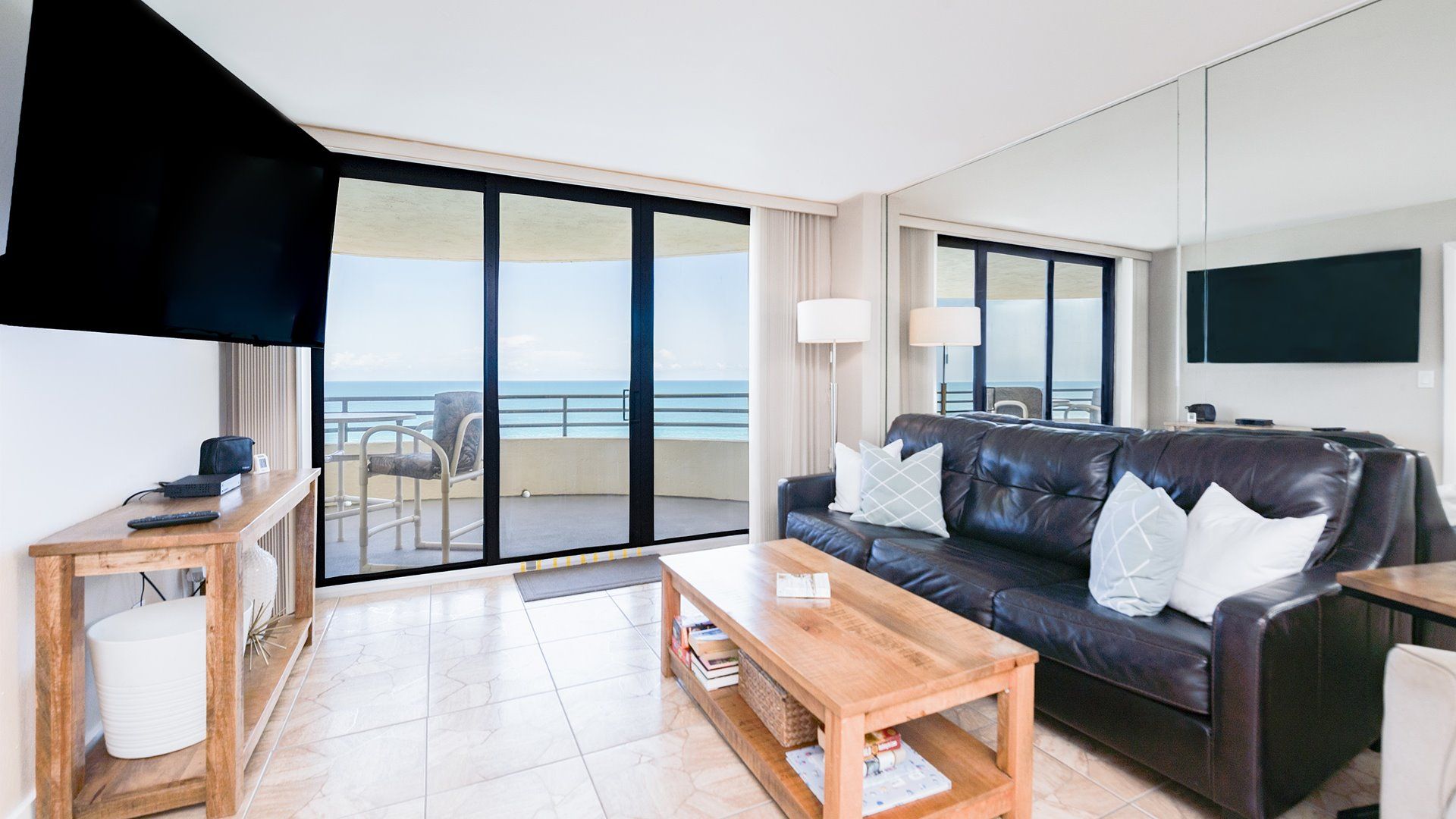 A hotel room with a balcony, a leather sofa, a wooden coffee table, a flat-screen TV, and a desk with a lamp.
