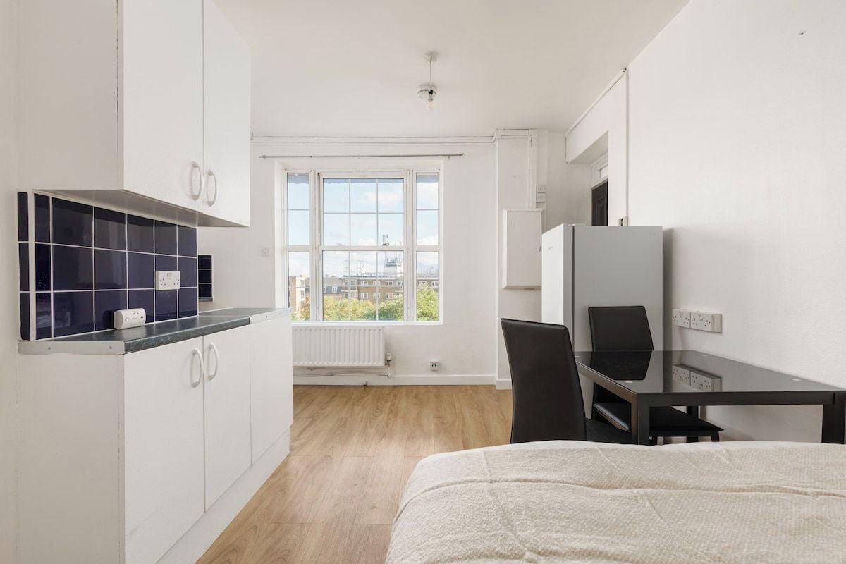 A modern hotel room with a bed, a desk, a chair, a kitchenette, and a large window offering a city view.