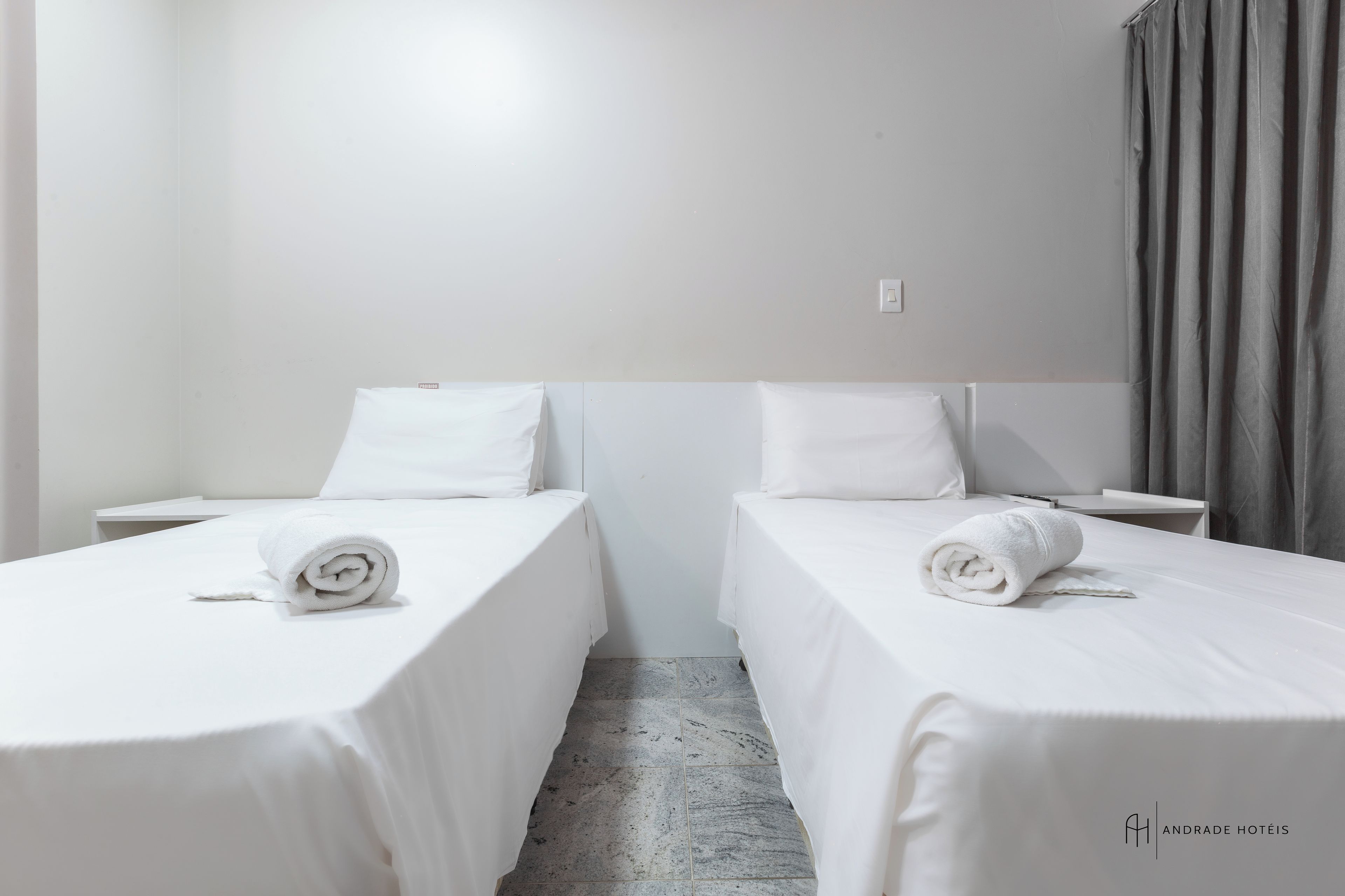 Une chambre d’hôtel avec deux lits simples, chacun agrémenté d’une serviette blanche soigneusement pliée, d’une petite table de chevet et d’un mur blanc.