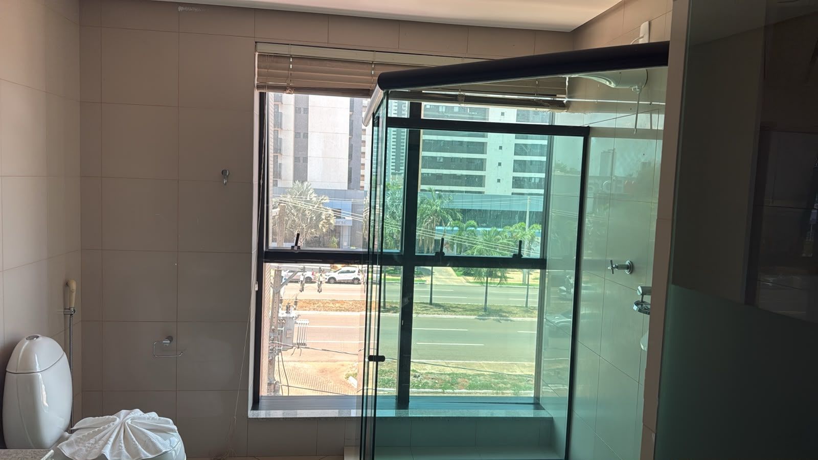 A bathroom with a glass shower enclosure, a toilet, and a window with a view of a street and buildings.