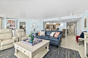 A spacious living room with a sofa, armchairs, and a coffee table. A bookshelf and a dining area are visible in the background. - #1404 Seychelles Oceanfront Traffic Free Beach (Daytona Beach Shores)
