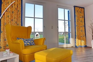 A room with a yellow armchair and ottoman, a white side table, and a view through a glass door to the outside. - Thatched house "Villa Selma" (Zierow)