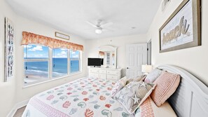 A bedroom with a large window, a ceiling fan, a television, and a bed with patterned bedding. - 1701 Towers Ten in Daytona Beach Shores, FL (Daytona Beach Shores)