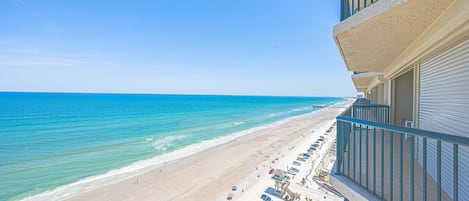 A beach view from a balcony with a clear sky, a sandy beach, and a calm sea.