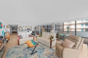 A spacious living area with a sofa, armchairs, and a coffee table. There is a dining area with a table and chairs, and a kitchen in the background. - 1B Royal Atlantic in Daytona Beach Shores, FL (Daytona Beach Shores)